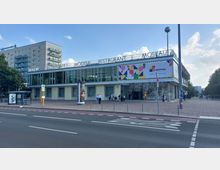 Modernes Gebäude mit der Aufschrift „Moskau“ und großen Glasfenstern in der oberen Etage, darunter ein Eingangsbereich mit Mosaikdekoration. Davor eine breite Straße mit Bushaltestelle und wenige Passanten auf einem gepflasterten Platz.