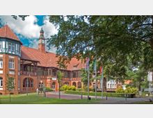 Backsteingebäude im Stil des Historismus mit roten Ziegeldächern, Fachwerkelementen und einem Turm mit Uhr. Im Vordergrund befinden sich Bäume, eine Rasenfläche, Gehwege und mehrere Flaggenmasten.