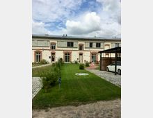 Ein modernisiertes Gebäude mit hellen Wänden und roten Backsteinakzenten, umgeben von einem gepflegten Rasen und gepflasterten Wegen. Im Vordergrund ist ein überdachter Stellplatz mit einem weißen Auto sichtbar, der Himmel ist bewölkt.