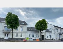 Straßenszene mit einer Reihe weißer historischer Gebäude mit Ornamentfassaden und dunklen Ziegeldächern. Zwei große Bäume stehen vor den Gebäuden; im Vordergrund ist ein Gehweg und eine Straße mit Verkehrszeichen erkennbar, bei bewölktem Himmel.