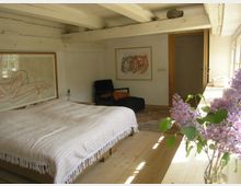 Schlafzimmer mit Holzbalkendecke und Holzboden, ausgestattet mit einem Bett mit gestreifter Tagesdecke, einem schwarzen Sessel, zwei Wandbildern und einem Teppich. Auf der Fensterbank im Vordergrund steht eine Vase mit lila Fliederblüten.