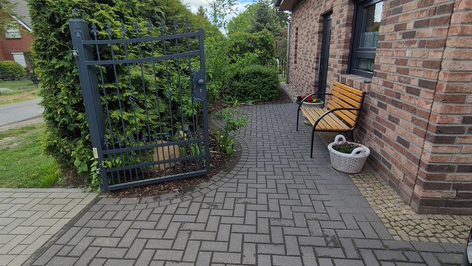Gepflasterter Eingangsweg neben einem Backsteinhaus mit Dachüberstand und Fenster, an der Wand steht eine Holzbank mit Pflanzkübel. Links befindet sich ein schwarzes Metalltor mit Hecken und Sträuchern entlang der Grundstücksgrenze.