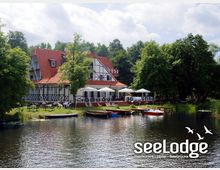 Fachwerkgebäude mit rotem Satteldach und weißer Fassade direkt am Ufer eines Sees, umgeben von Bäumen. Vor dem Gebäude befinden sich Terrassen mit Tischen, Sonnenschirmen und kleinen Booten am Wasser.
