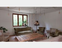 Wohnzimmer mit hellen Wänden, Holzboden und einer Sitzgruppe aus grünen Polstermöbeln sowie gestreiften Sesseln. Im Raum hängen mehrere Glühbirnen an Kabeln von der Decke, ein rustikaler Holztisch steht zentral, und eine Stehlampe sowie Pflanzen befinden sich vor einem Fenster mit Holzrahmen, das nach draußen ins Grüne blickt.