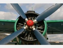 Nahaufnahme eines Flugzeugpropellers mit vier blauen Rotorblättern und einer roten Nabe, umgeben von einem grünen Gehäuse. Im Hintergrund sind Teile des Flugzeugflügels, ein bewölkter Himmel und Gebäude zu sehen.
