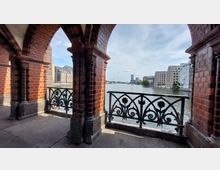 Blick durch die roten Backsteinbögen der Oberbaumbrücke in Berlin auf die Spree, mit kunstvoll verzierten Eisen-Geländern im Vordergrund und modernen Gebäuden entlang des Flusses im Hintergrund. Der Himmel ist leicht bewölkt.