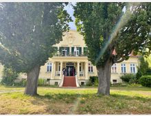 Helles, herrschaftliches Landhaus mit Säulenportal, breiter Freitreppe und Balkon mit grünen Fensterläden, vor der Fassade ein Wappenrelief. Das Gebäude liegt in einem parkartigen Garten mit großen Bäumen, Rasenfläche und Sträuchern.