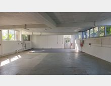 Leerer, rechteckiger Raum mit weißen Wänden, einer abgehängten Decke und großen Fenstern, die auf zwei Seiten Tageslicht hereinlassen. Am hinteren Ende befindet sich eine Glastür, daneben Feuermelder und Feuerlöscher an der Wand befestigt.