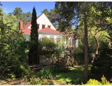 Ein weißes Haus mit rotem Satteldach, umgeben von einem üppig bewachsenen Garten mit hohen Bäumen, Sträuchern und Blumen. Im Vordergrund ist ein gepflasterter Weg und Steinmauern zu sehen, während der Hintergrund von Wald umgeben ist.