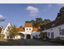 Hof eines historischen Gutshofs mit weiß verputzten Gebäuden, roten Ziegeldächern und arkadenartigem Torbogen. Im Hintergrund Wald und blauer Himmel, im Vordergrund Bänke und Kiesboden.
