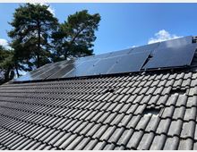 Dach eines Hauses mit schwarzen Ziegeln, auf dem mehrere Solarpaneele montiert sind. Im Hintergrund stehen grüne Nadelbäume vor einem blauen Himmel.