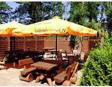 Außenbereich eines Biergartens mit Holzbänken und -tischen sowie orangefarbenen Sonnenschirmen der Marke "Schöfferhofer Weizen". Im Hintergrund sind Holzzäune, grüne Bäume und eine Laterne zu sehen.