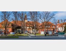 Backsteinbau mit roten Ziegeldächern und großen weißen Fenstern, umgeben von kahlen Bäumen und Grünflächen, im Vordergrund eine Straße mit Verkehrsschildern, Laternen und einer Bushaltestelle. Im Hintergrund ragt ein Turm mit einer grünen Kupferspitze in den Himmel.