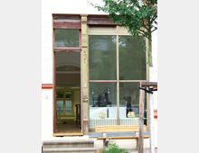 Fassade eines historischen Gebäudes mit großer Fensterfront und geschnitzten Holzdetails. Hinter dem Fenster sind Schreibtischlampen, ein Heizkörper und Brötchen auf einer Ablage sichtbar; im Vordergrund stehen eine Holzbank und ein Baum.