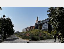 Eine moderne städtische Szenerie mit einem Gehweg, der von Bäumen gesäumt ist, und einem Hügel mit Gras in der Mitte, hinter einem Zaun. Im Hintergrund sind moderne Gebäude mit großen Glasfassaden und ein hoher Schornstein zu sehen, unter einem klaren blauen Himmel.