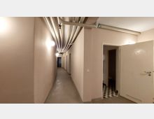 Langer, schlichter Korridor mit beige gestrichenen Wänden und Deckenrohren an der Oberseite. Auf der rechten Seite befindet sich eine offene Tür, die zu einem Raum mit schwarz-weiß kariertem Boden führt.