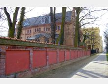 Backsteinbau im neugotischen Stil mit hohen Fenstern und einem Ziegeldach im Hintergrund, flankiert von einer roten Backsteinmauer mit eingelassenen Paneelen und einem Gehweg im Vordergrund. Die Szene ist von kahlen Bäumen gesäumt.