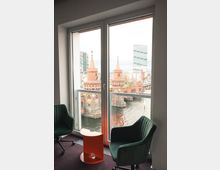 Blick aus einem modernen Raum mit zwei grünen Sesseln und einem kleinen orangen Beistelltisch auf die Oberbaumbrücke in Berlin, die über die Spree führt, umrahmt von hoher moderner und historischer Architektur.