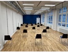 Ein heller Raum mit Holzboden, weiß gestrichenen Wänden und großen Fenstern auf der rechten Seite. Mehrere schwarze Stühle sind in gleichmäßigen Abständen aufgestellt; an der Stirnwand in Blau befindet sich eine Tür.