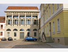 Historisches Gebäudeensemble mit gepflastertem Platz im Vordergrund; in der Mitte ein helles Gebäude mit Säulenfassade und roten Dachziegeln, daneben ein gelbes Wohnhaus mit Treppe und schmiedeeisernem Geländer. Mehrere geparkte Autos stehen vor den Gebäuden.