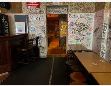 Innenraum einer alternativen Kneipe mit eng gestellten Holztischen und Hockern sowie einer dunklen Theke links. Die Wände und der Durchgang in den hinteren Raum sind dicht mit Graffiti und Zeichnungen bedeckt, darunter ein großes Totenkopf-Motiv.