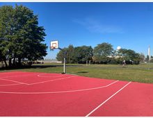 Ein rotes Basketballfeld im Freien mit einem einzelnen Basketballkorb; im Hintergrund eine Wiese, mehrere Bäume und eine Person auf einem Fahrrad. Am Horizont sind ein Gebäude und eine Radarkuppel sichtbar unter einem blauen Himmel.