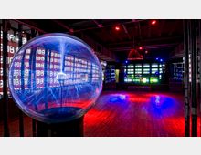 Ein futuristischer Innenraum mit einer großen Plasmakugel im Vordergrund, umgeben von leuchtenden Lichtblitzen. Im Hintergrund befinden sich mehrere Bildschirme mit Zahlenmustern und bunte Beleuchtung, die eine technologische und moderne Atmosphäre schaffen.