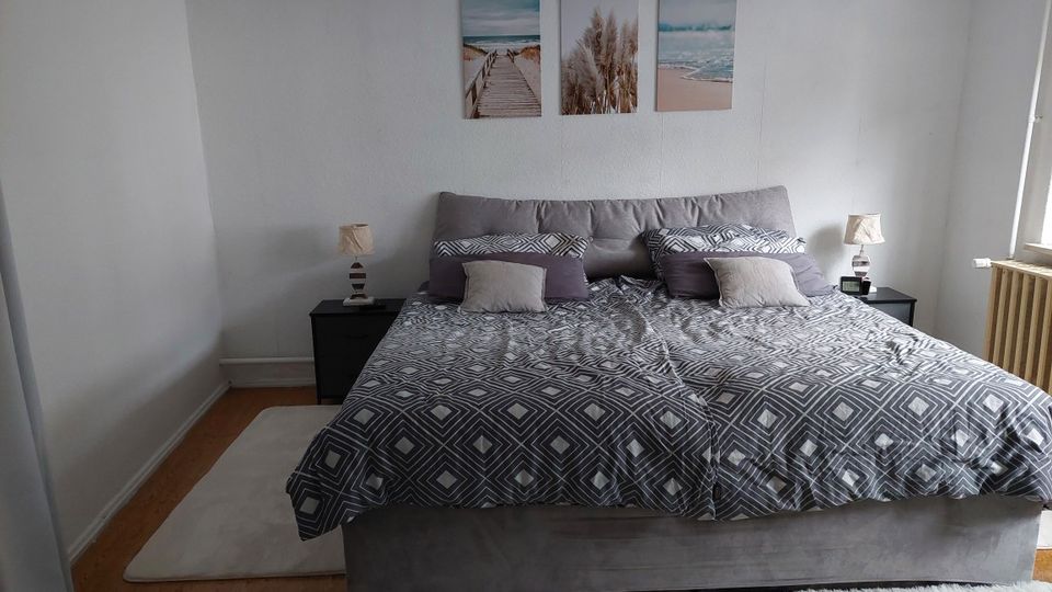 Schlichtes Schlafzimmer mit großem Doppelbett in grauer Bettwäsche, zwei Nachttischen mit Lampen und drei Wandbildern über dem Kopfteil. Rechts ein Fenster mit Heizkörper darunter, Holzboden und heller Teppich vor dem Bett.