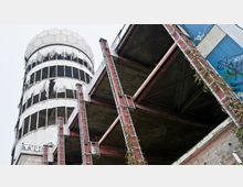 Eine verlassene Radarkuppel und ein Gebäudeteil einer ehemaligen Abhörstation mit freiliegenden Metallstreben, teilweise eingestürztem Dach sowie Graffitis an den Wänden. Die weiße Kuppel ist mit zerrissenem Material bedeckt, und Pflanzen ranken an den Metallstrukturen empor.