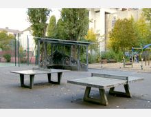 Öffentlicher Parkbereich mit Tischtennisplatten aus Beton, einem überdachten Sitzbereich aus Metall und einem angrenzenden Spielplatz mit Klettergerüst und Sandboden. Im Hintergrund sind Bäume, eine Umzäunung und Gebäude sichtbar.