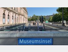 Eingang zur U-Bahn-Station "Museumsinsel" mit Rolltreppen und Treppen, flankiert von einem historischen, rosafarbenen Gebäude mit verzierten Fenstern auf der linken Seite. Im Hintergrund befinden sich Bäume und weitere Gebäude.