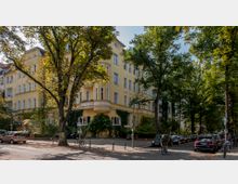 Eckhaus in einer städtischen Wohngegend mit gelber Fassade, großen Fenstern und einem verzierten Balkon. Umgeben von Bäumen, parkenden Autos und vereinzelten Fahrrädern, vermittelt die Straße eine ruhige Umgebung mit viel Grün.