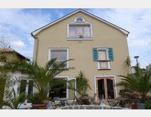 Beigefarbenes Haus mit zwei Etagen und Satteldach, großen Fenstern und blau gestrichenen Fensterläden auf einer Fensterseite. Im Vordergrund befinden sich eine Terrasse mit Sitzmöbeln und mehrere in Töpfen gepflanzte Palmen.