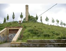 Ein steiler, mit Gras bedeckter Hügel mit einem hohen, sechseckigen Ziegelturm an der Spitze. Im Vordergrund ist ein mit Graffiti bedeckter Eingang zu einem gemauerten Durchgang sichtbar.