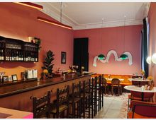 Gemütliche Bar mit rosa Wänden und einem langen Holztresen mit hohen Holzstühlen. Im Hintergrund befinden sich Sitzbänke mit orangefarbenem Polster, runde Marmortische und grüne Hängelampen; dekorative Wandkunst und Pflanzen unterstreichen das stilvolle Ambiente.