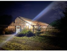 Ein beleuchtetes Gewächshaus bei Nacht, mit Glaswänden und einem spitz zulaufenden Dach. Im Vordergrund sind Büsche und Sträucher, während rechts ein weiterer Gewächsrahmen zu sehen ist. Ein heller Lichtstrahl scheint auf das Gelände.