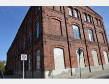 Backstein-Gebäude mit historischer Fassade, bestehend aus symmetrischen Rundbogenfenstern und dekorativen Zierelementen. Im Vordergrund ein Verkehrsschild, das eine Tempo-30-Zone anzeigt, und ein gepflasterter Gehweg entlang der Gebäudewand.