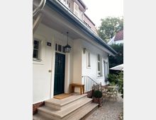 Weiße Hausfassade mit grüner Eingangstür und einer Treppe mit Steinfliesen. Vor dem Eingang stehen ein Holzbank, ein kleiner Tisch mit Stühlen und Pflanzen in Töpfen; im Hintergrund sind Dachgauben und Bäume sichtbar.