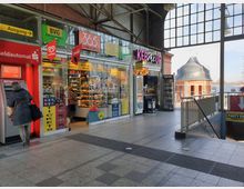 Bahnhofshalle mit Geschäften und Geldautomat, darunter ein Kiosk mit Snacks, Getränken und Tickets sowie ein Imbiss namens „Keb Me Up“. Im Hintergrund sind große Fenster mit Blick nach draußen und ein rundes Backsteingebäude sichtbar.