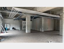 Rohbau eines Innenraums mit sichtbaren Betonwänden, freiliegenden Lüftungsrohren und Kabeln an der Decke. Der Boden ist mit Fliesen bedeckt, und Baumaterialien sind am Rand verteilt.