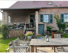 Backstein-Gebäude mit rotem Ziegeldach, vorgelagertem Holzbalkon und angrenzendem Garten. Im Vordergrund befinden sich Gartenmöbel aus Holz, Blumentöpfe und eine Laterne auf einem großen Holztisch; Holzscheite sind unterhalb des Balkons gestapelt.