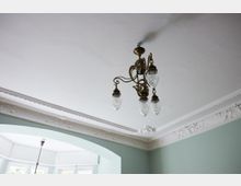 Ein Innenraum mit weißer Decke, einem dekorativen Kronleuchter aus Metall mit gläsernen Lampenschirmen und verzierten Stuckleisten. Die Wände sind in einem hellgrünen Farbton gestrichen, und ein großer, bogenförmiger Fensterbereich mit weißen Holzrahmen lässt Tageslicht herein.