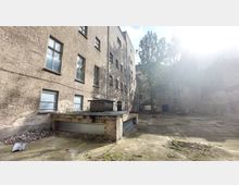 Hinterhof eines alten Mehrfamilienhauses mit hellbrauner Fassade und sichtbaren Abnutzungsspuren. Der Hof ist größtenteils betoniert, mit einzelnen Pflanzen und einem Baum in der Mitte, umgeben von hohen Wänden und vereinzelten Graffitis.