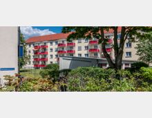 Mehrstöckiges Wohngebäude mit beigen Fassaden, roten Balkonen und einem roten Satteldach, umgeben von Bäumen, Sträuchern und einem begrünten Innenhofbereich. Im Vordergrund befindet sich ein Schild mit der Aufschrift „Kastanienweg 9, 11, 13“.