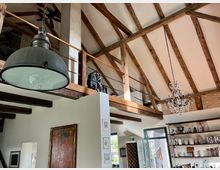 Innenraum eines rustikalen, modernen Wohnbereichs mit hohen Decken und sichtbaren Holzbalken. Zu sehen sind ein Kronleuchter, eine große Hängelampe, Regale mit Büchern und Deko sowie eine kleine Galerie mit einem Geländer aus Holz und Metallketten.