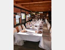 Langer, festlich gedeckter Tisch in einem rustikalen Raum mit Backsteinwänden und Holzdecke, dekoriert mit weißen Stuhlhussen, Kerzen und Blumenarrangements; im Hintergrund sind Metalltanks und kupferne Destillationseinheiten sichtbar.