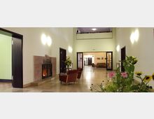 Ein heller Eingangsbereich mit großen Fliesen, einem offenen Kamin auf der linken Seite, Sitzmöbeln und Blumenarrangements. Im Hintergrund sind eine Glasdoppeltür und weitere Räume sichtbar.