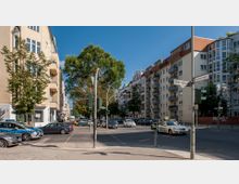 Straßenecke in einer Stadt mit mehrstöckigen Wohngebäuden auf beiden Seiten, Ampeln, Straßenschildern (Steifensandstraße und Suarezstraße) und mehreren geparkten sowie fahrenden Autos, darunter ein Taxi und ein Polizeifahrzeug. Bäume säumen die Straße unter einem blauen Himmel.