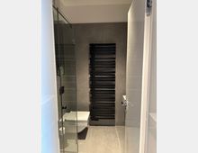 Modernes, grau gefliestes Badezimmer mit einer Glasdusche links und einem wandhängenden WC. An der Rückwand befindet sich ein großer, schwarzer Handtuchheizkörper mit horizontalen Streben.