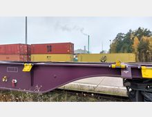 Ein Güterbahnhof mit gestapelten gelben und roten Containern im Hintergrund. Im Vordergrund ist ein violettes Bahnwaggongestell, und rechts sind Herbstbäume sowie eine Industrieanlage mit einem rauchenden Schornstein sichtbar.